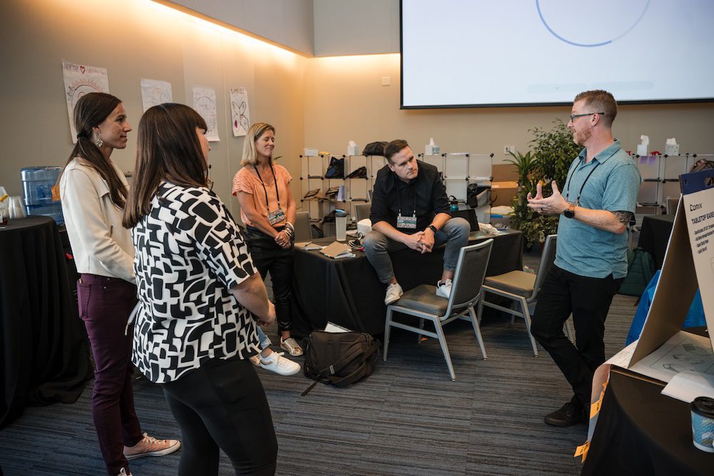 A group of five people are engaged in a discussion in a conference room. One person is speaking while the others listen. The room has conference materials, a projector screen, and chairs. Some posters are visible on the walls.