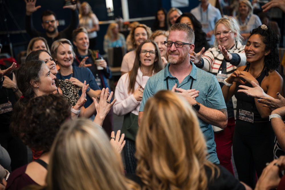 A group of people stands in a circle around a man with glasses who has his hand on his chest. They appear to be sharing an interactive or participatory moment indoors, with many smiling and engaged.