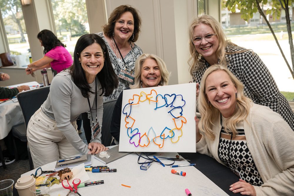 Five people are smiling and holding a canvas with a design of interconnected colorful hearts. They are seated around a table with art supplies. The room has windows showing trees outside.