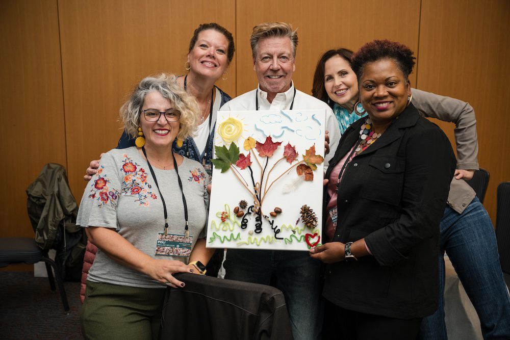 A group of five people smiling, holding a framed artwork featuring leaves, twigs, and other natural elements. They are indoors, standing against a wood-paneled wall.