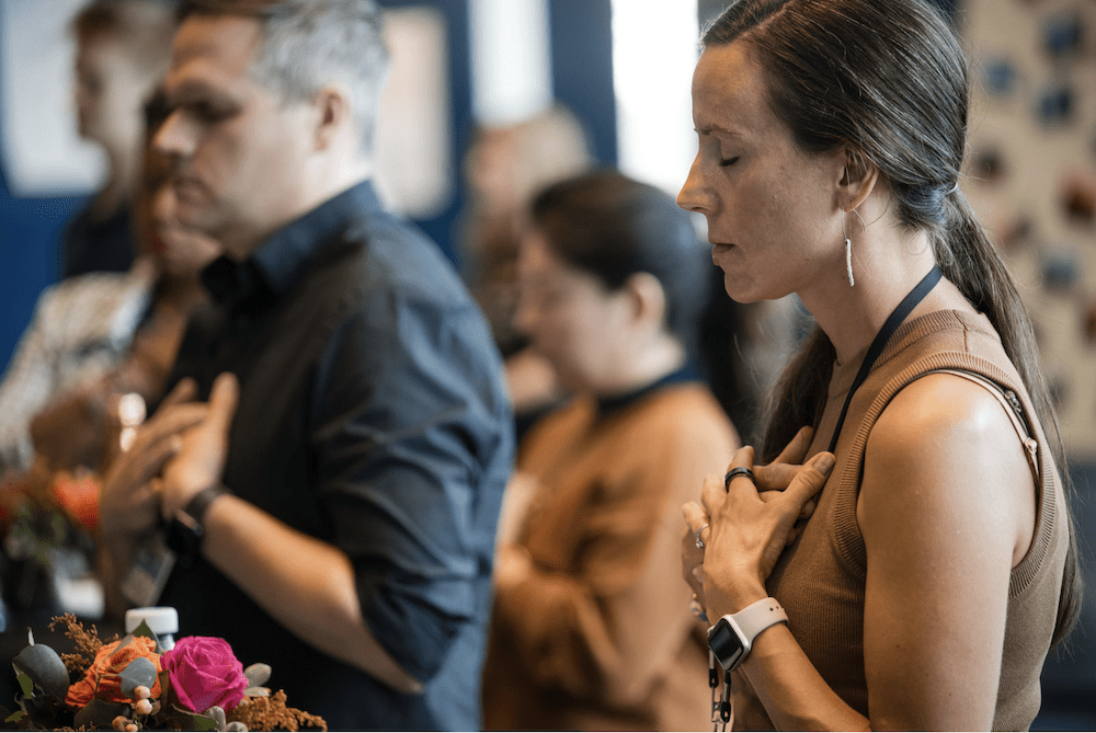 A group of people stand with their eyes closed and hands over their hearts, appearing to be in a moment of reflection. The individuals are in a room with blurred background elements, including flowers on a table in the foreground.