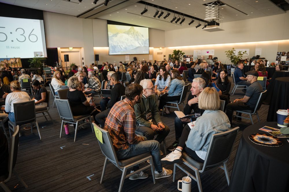 A large group of people sits in clusters in a conference room with a high ceiling. A screen displaying a mountain image is at the front. Attendees are engaged in discussions. Tables and chairs are arranged throughout the room.