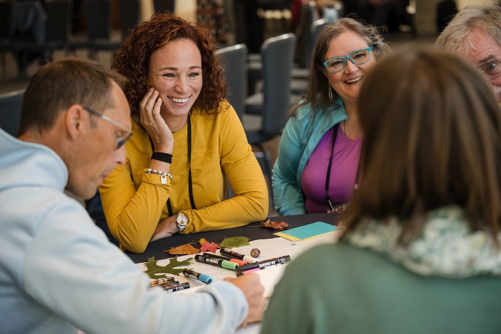 Four people sit around a table engaged in discussion. The woman with curly hair in a yellow shirt smiles, listening to a man writing on paper. Others, including a woman with glasses, also participate. Pens and leaves are on the table.