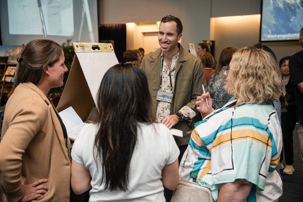 A group of people engaged in conversation near an easel at a conference or event. A man stands facing three women, smiling and discussing. Attendees in the background with screens and presentations visible.