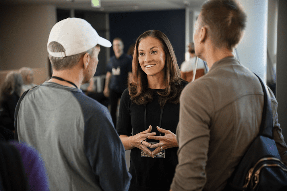 A woman smiles and gestures with her hands while talking to two men in a casual setting. One man wears a white cap and a gray shirt, and the other wears a khaki jacket and carries a bag over his shoulder. People are in the background.