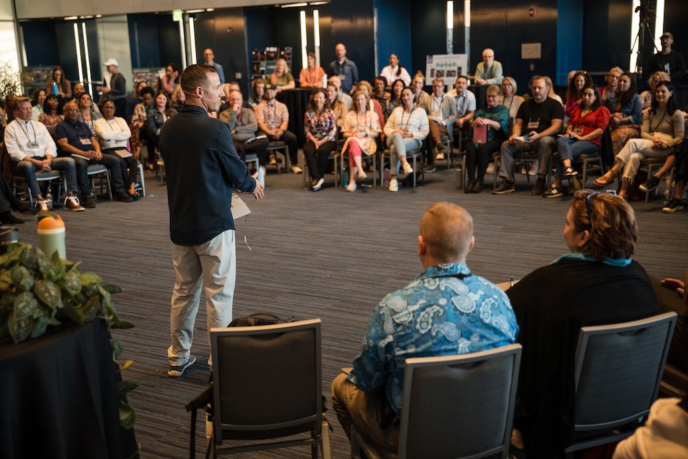 A man stands speaking to a large group of seated people in a conference room. The audience forms a semi-circle around him, with two people seated in the foreground. The room has dark blue walls and natural lighting.