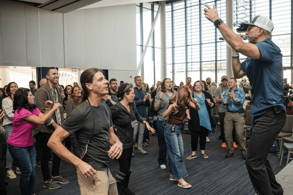 A group of people stand and engage with an energetic speaker at the front of a spacious room with large windows. Some are clapping or gesturing, while others smile and pay attention. The setting appears to be a presentation or workshop.