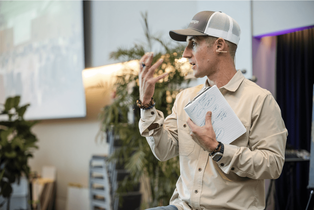 A person wearing a beige shirt and cap is speaking and gesturing with one hand while holding a notebook in the other. The background features a plant and a projection screen.