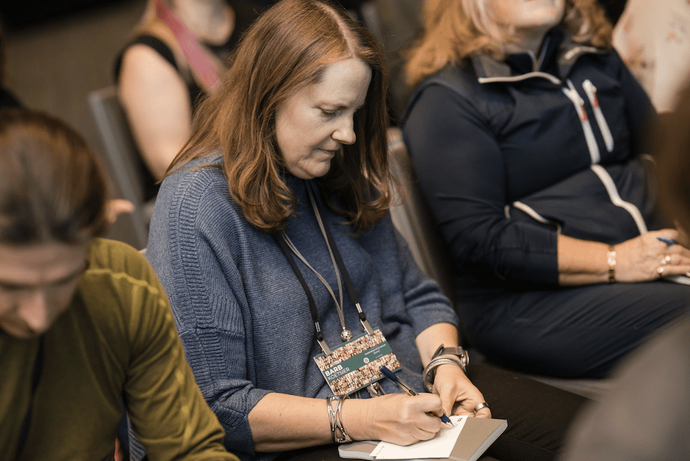 A woman with long brown hair sits in an audience taking notes on a notepad. She is wearing a blue sweater and a conference badge. Other people are seated around her, some holding pens. The setting appears to be a conference or seminar.