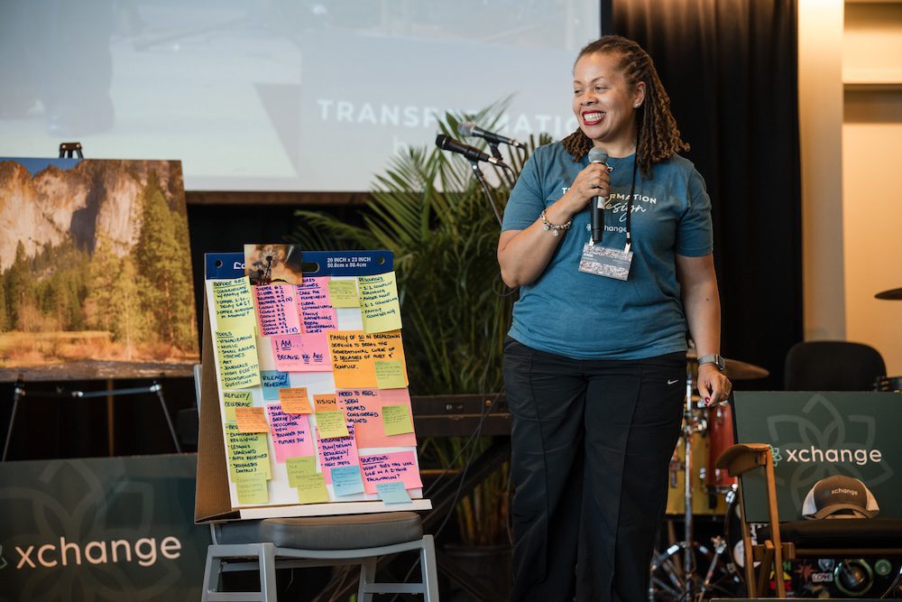 A person stands on a stage with a microphone, smiling, next to a poster board covered in colorful sticky notes. There is a large painting of a landscape to the left and a drum set partially visible in the background.