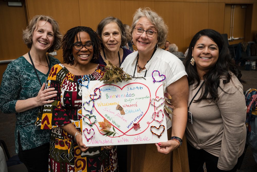 A group of five people stand indoors, smiling at the camera. They hold a colorful sign with the word "Welcome" written in multiple languages, surrounded by hearts and decorative leaves. The background features a wood-paneled wall.