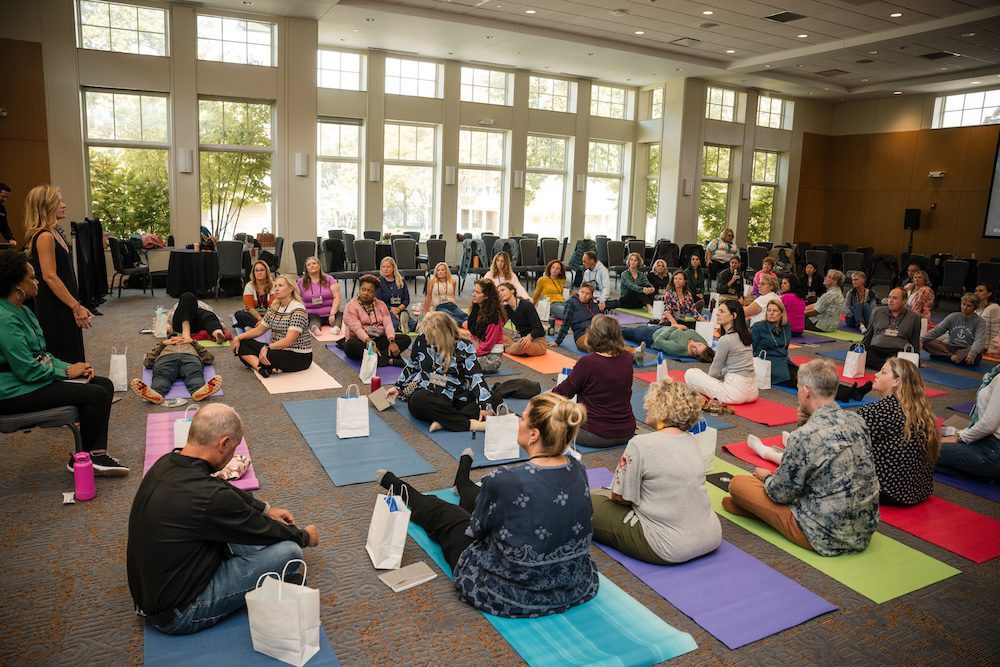 A group of people participating in a workshop are seated on colorful yoga mats in a large conference room. Some people are lying down while others are sitting upright. Large windows in the background let in natural light.