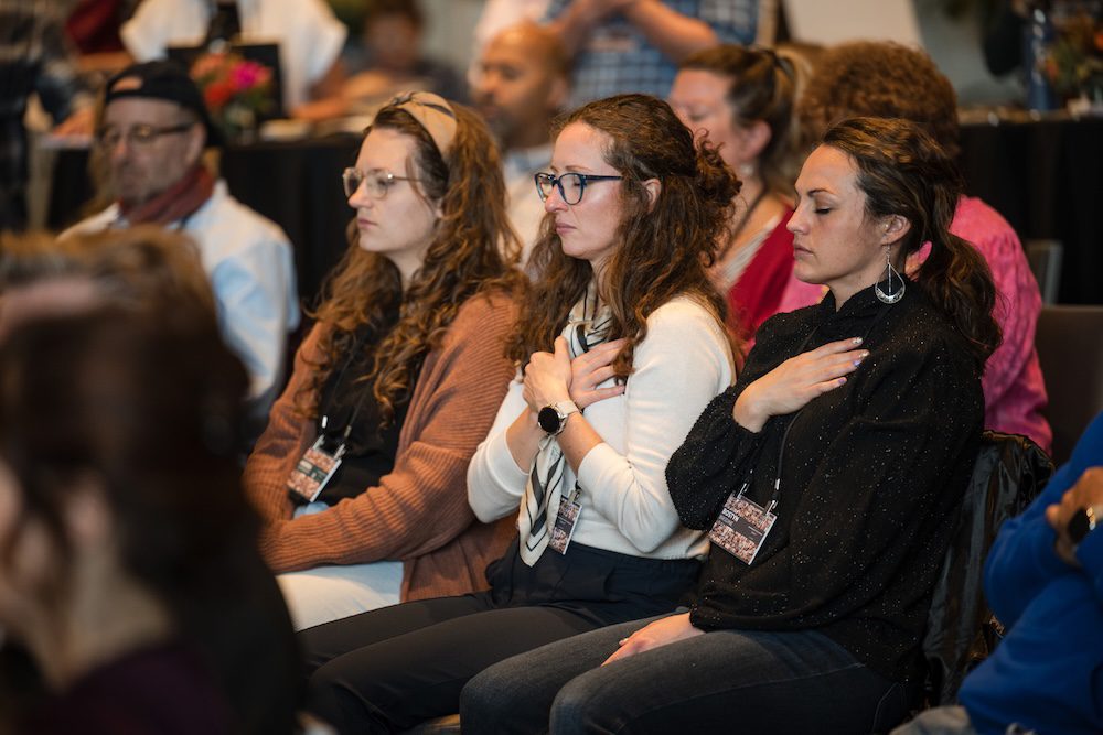 A group of people sit in a row during an event, each with their eyes closed and a hand placed over their chest. They appear focused and are wearing name tags. The setting suggests a conference or seminar environment.