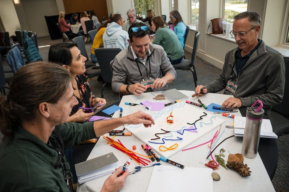 A group of people sitting around a table engaged in an art activity. They are using colorful markers, pipe cleaners, and papers. The setting appears to be a workshop or meeting room with others in the background.