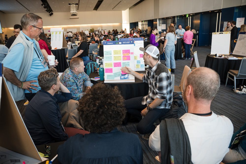 A group of people are gathered around a display board covered with colorful sticky notes. Some are seated, and one person is kneeling, pointing to the board. The setting appears to be a busy conference or workshop in a large room.