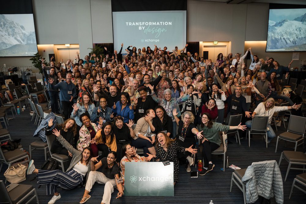 A large group of people pose energetically in a conference room with two projection screens displaying "TRANSFORMATION BY design." They are surrounded by chairs and a sign on the floor reads "xChange.