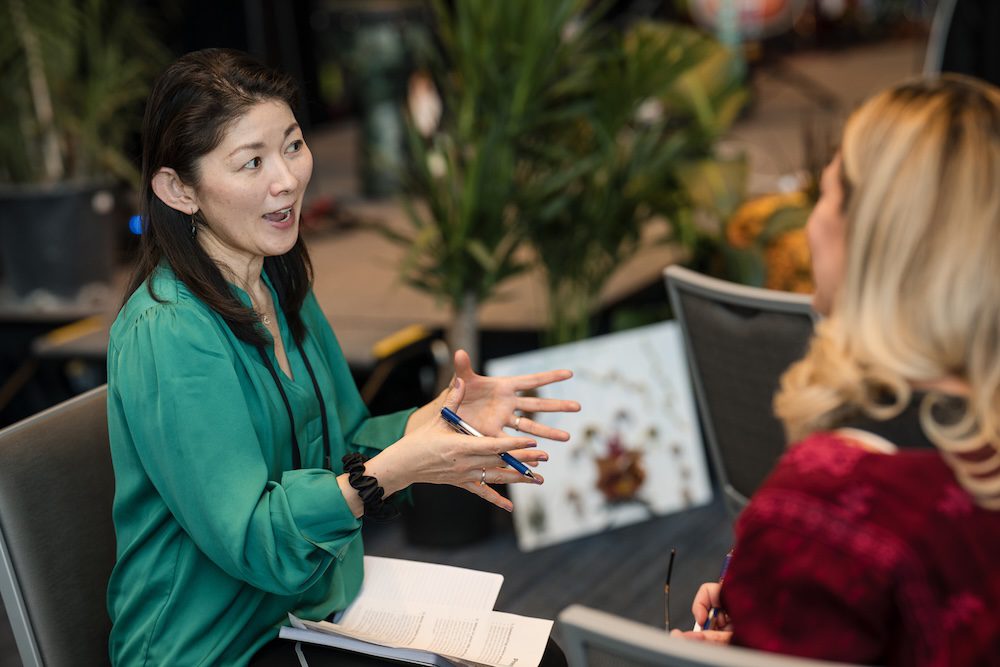 Two women are sitting in chairs engaged in conversation. The woman on the left is gesturing with her hands while holding a pen. There are plants in the background. A notepad is visible in her lap.