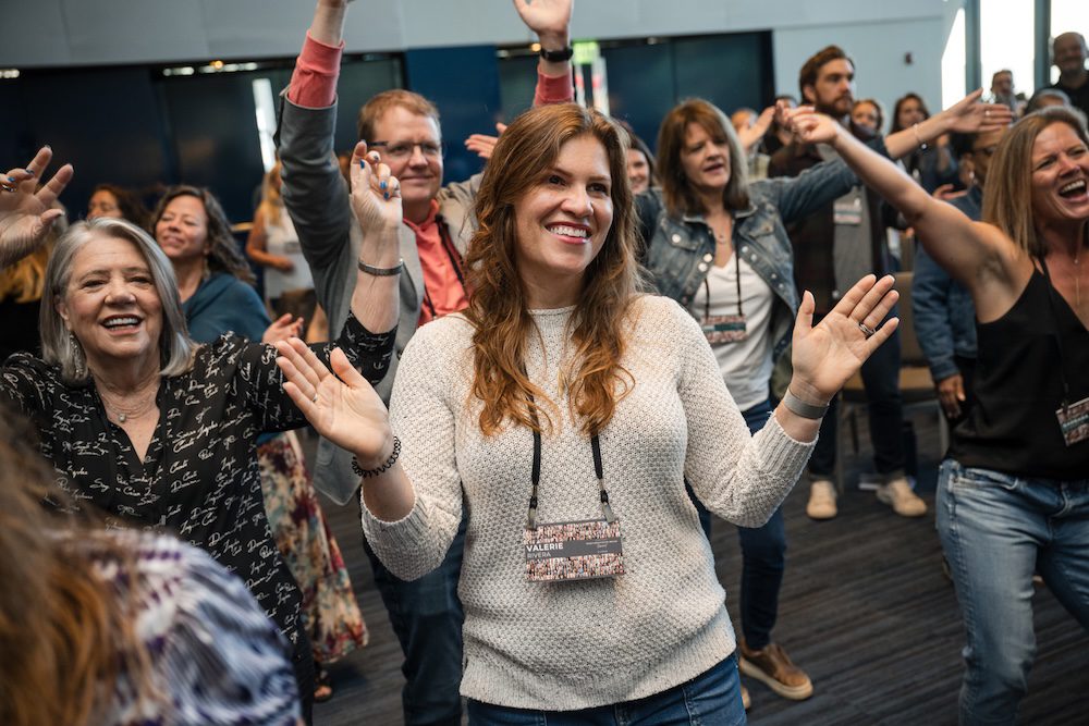 A group of people stands in a room, appearing to dance or celebrate. They are smiling and have their hands raised. They wear casual clothing and name tags. The setting appears to be a conference or social event.