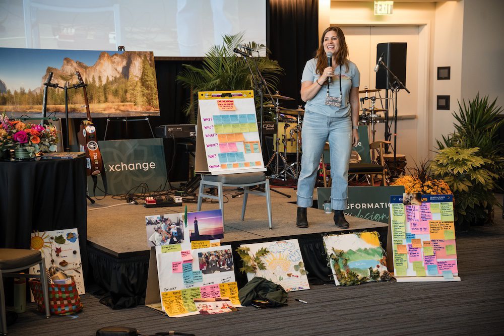 A woman stands on stage holding a microphone at a presentation. The stage features various colorful posters and a painting of a landscape. Musical instruments are visible in the background, and a banner with the word "exchange" is partially shown.