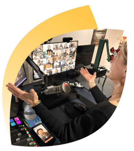 A person sits at a desk engaged in a Facilitation Masterclass video call with multiple participants displayed on two monitors. The desk is equipped with a microphone, audio mixer, and smartphone, ready to create learning experiences. Arms are outstretched in a welcoming gesture.