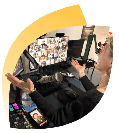 A person with light hair is sitting at a desk with multiple computer screens displaying a video conference. They have outstretched arms and a microphone in front of them. Audio equipment and a smartphone are also on the desk.